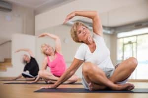 Active senior lady with group of women doing relaxing stretching workout for body flexibility in yoga studio.