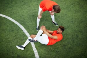 Injured soccer player on ground holding knee while team mate checks on him